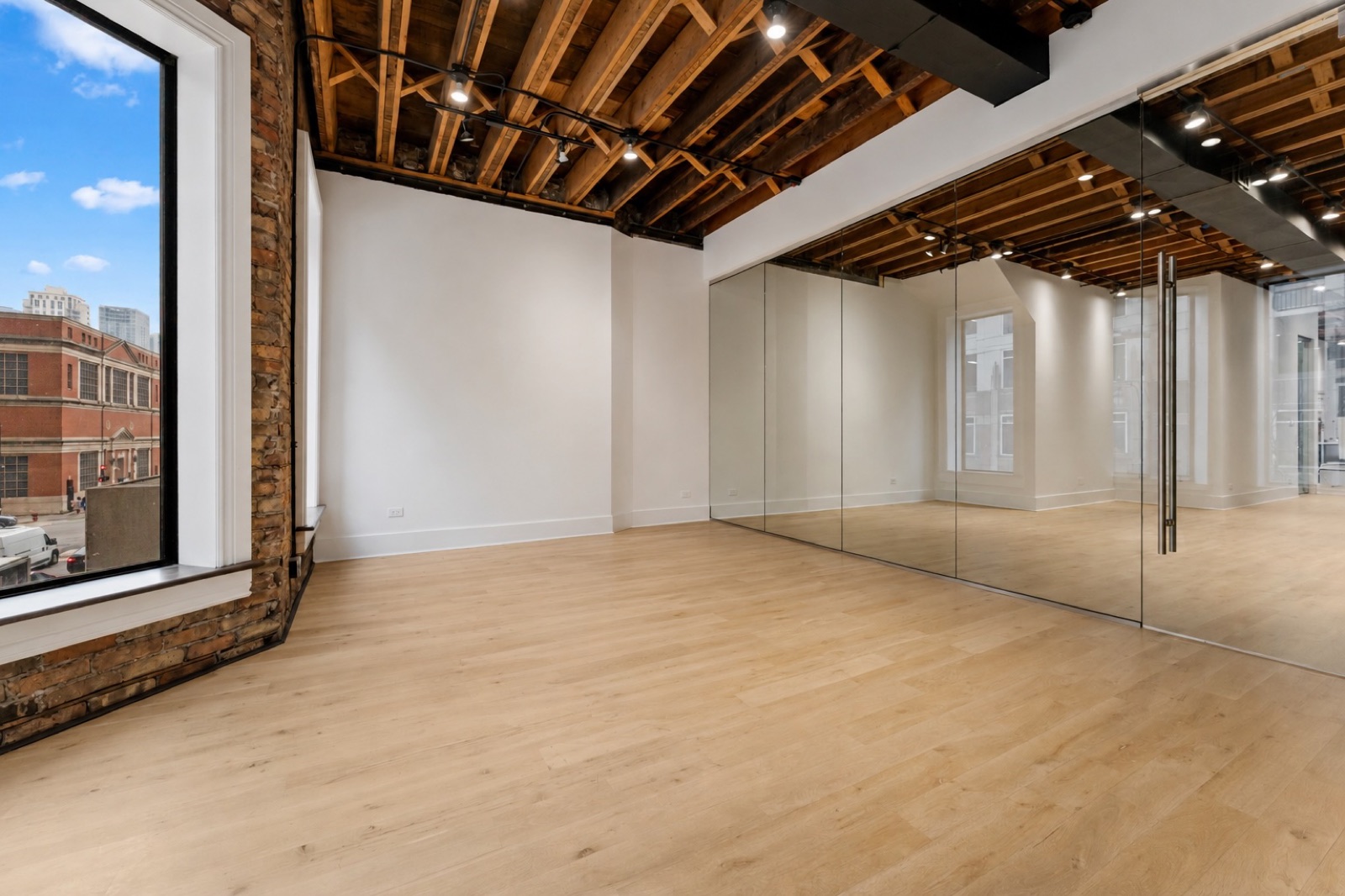 Loft A — bay window with city view, exposed brick, mirrored wall, and original wood-beam ceiling.
