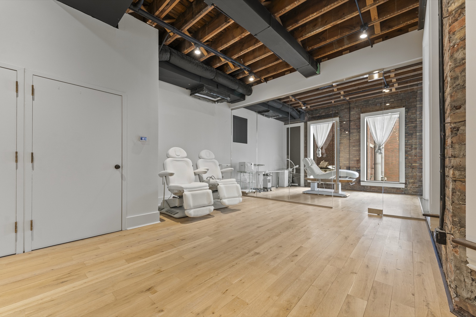 Loft C — compact studio with exposed brick, mirrored wall, wood-beam ceiling, and bay windows with sheer curtains.